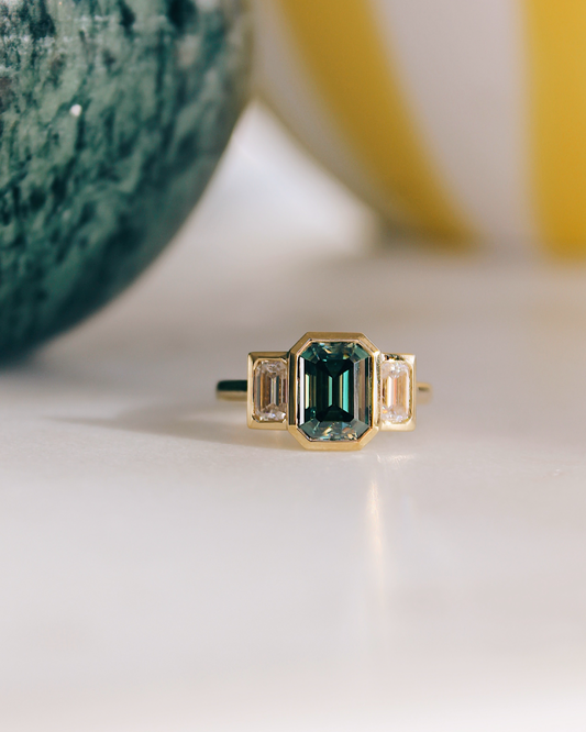 18K Trilogie Goldring Olivia mit einem grünen Emerald Cut Moissaniten und zwei Baguette Seitensteinen.